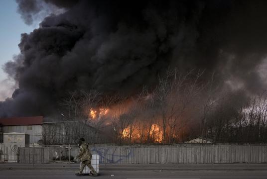 မတ် ၁၇ ရက်က ကိယက်ဗ်မြို့ ဆင်ခြေဖုံးဒေသတွင် ဗုံးကြဲခံရပြီးနောက် ဂိုထောင်တစ်လုံး မီးလောင်နေစဉ် ဝန်ထမ်းတစ်ဦး ကုန်ထုပ်တစ်ခုကို သယ်ယူသွားပုံ (Photo:AP)